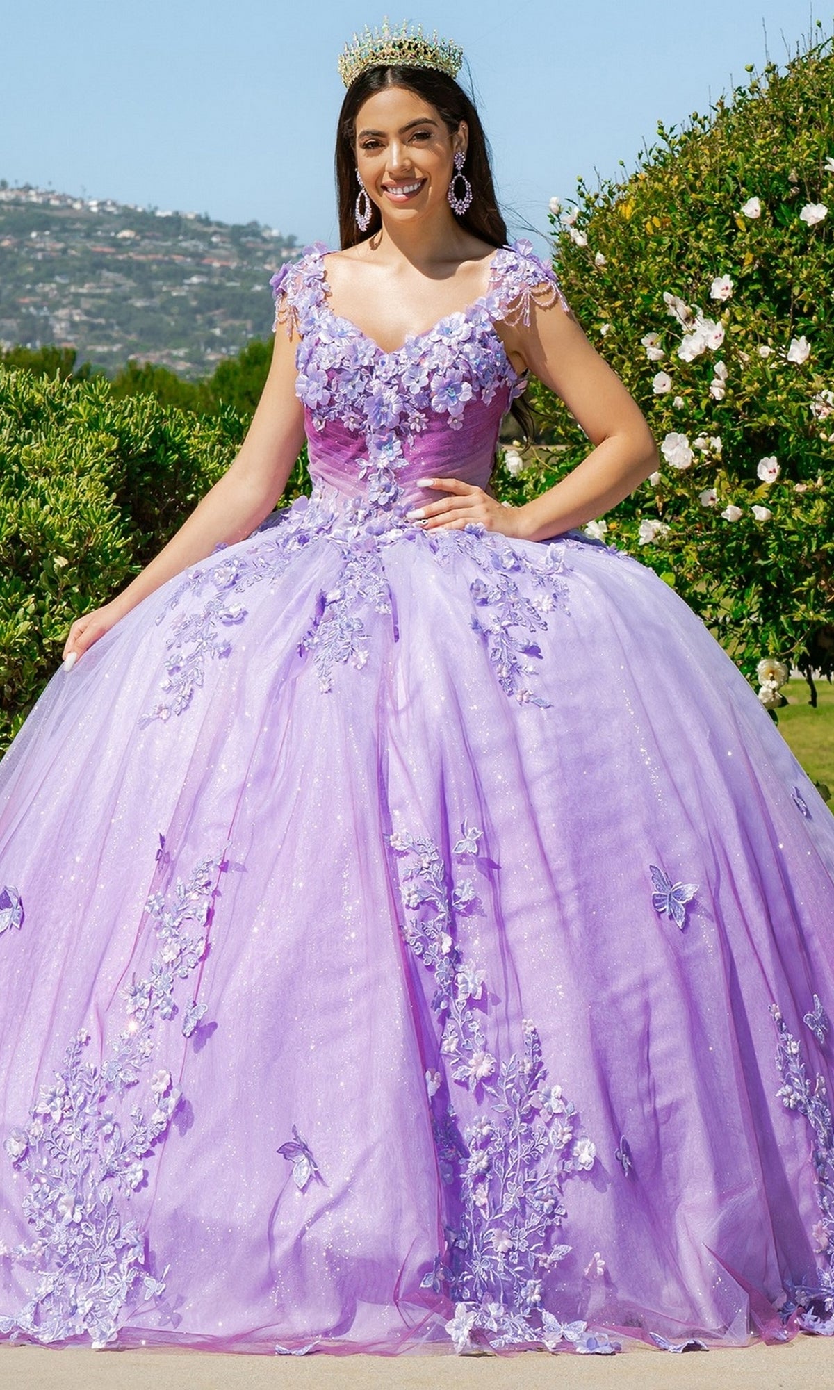 A woman wears the Quinceanera Ball Gown 8088J, featuring lavender fabric with 3D floral lace and butterfly details. She stands outdoors, smiling and wearing a tiara.